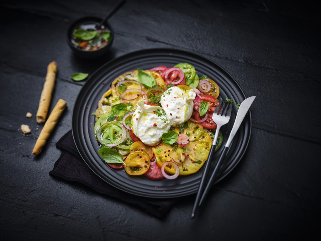 Heirloom tomato carpaccio topped with fresh Maestrella Burrata, red onion, basil and mixed herbs, plated on a dark dish for a professional foodservice presentation.