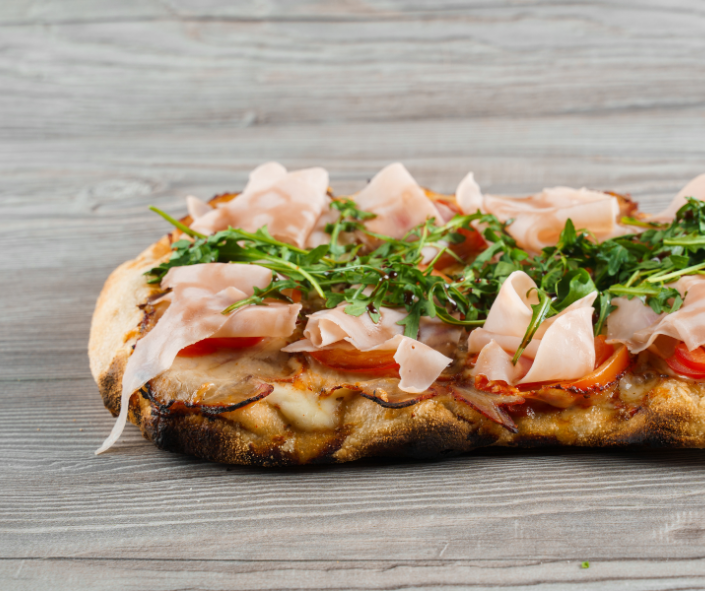Gourmet Pinsa Romana with melted mozzarella, Parma ham, fresh tomatoes and rocket on a charred light crust, illustrating premium topping versatility and consistent cheese performance for upscale professional foodservice menus.