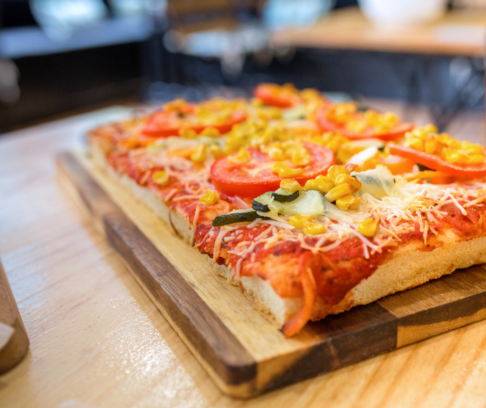 Rectangular al taglio style pizza topped with grated mozzarella, fresh tomatoes, corn and mixed vegetables on a wooden board, ideal for premium snacking and high-volume foodservice operations.