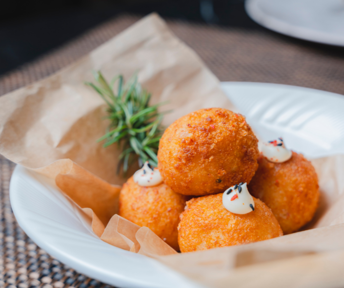 Golden fried mozzarella croquettes served in a white bowl with parchment paper, topped with a seasoned sauce and garnished with fresh rosemary for an elevated professional foodservice presentation.